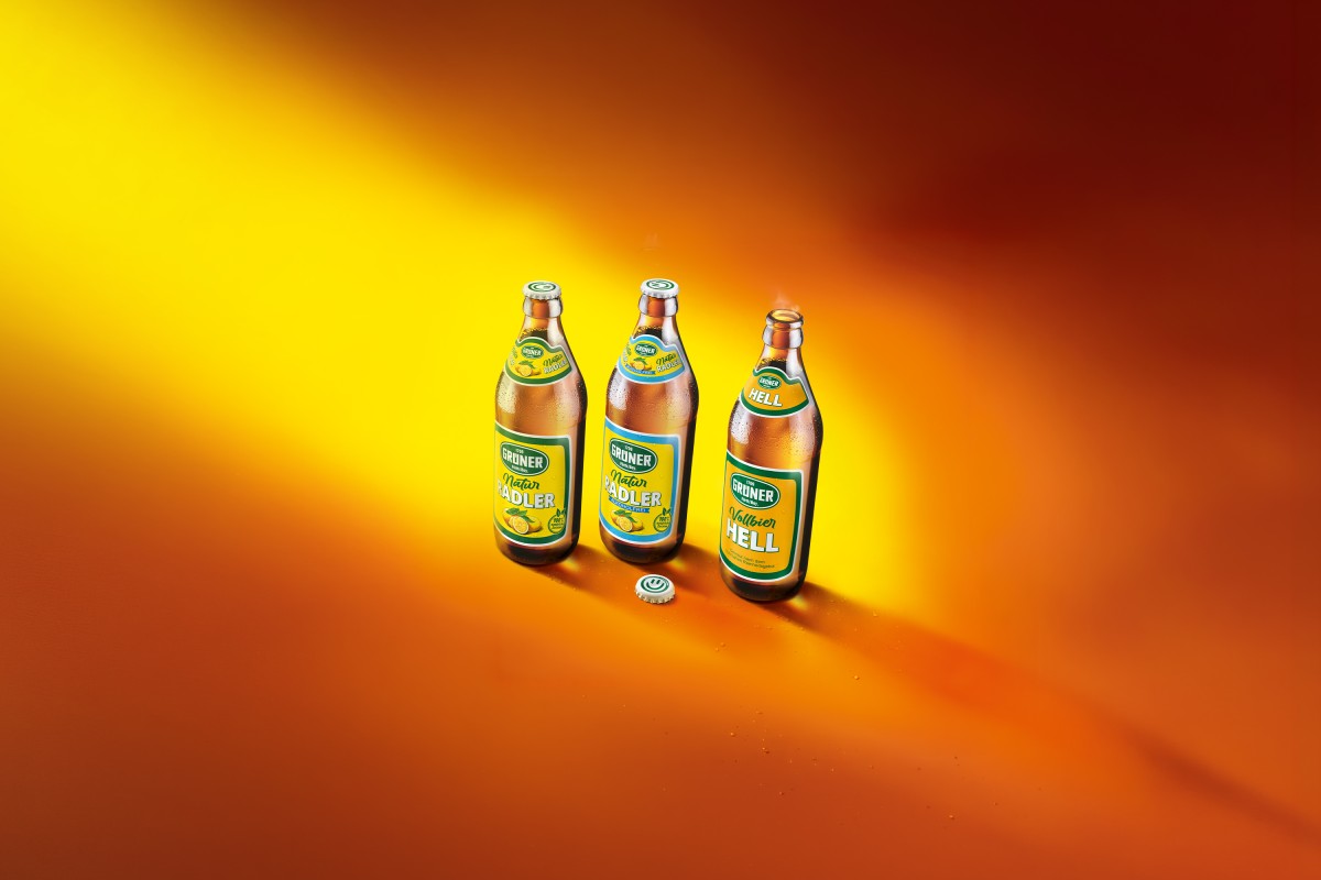 Drei Flaschen Grüner Bier der Sorte Vollbier Hell, Natur Radler und Natur Radler Alkoholfrei vor gelb-orangenem Hintergrund.